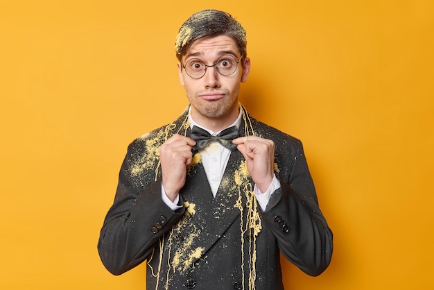 Surprised funny man adjustes bowtie dressed in formal outfit wears round spectacles dresses for special occasion smeared with serpantine spray isolated over yellow background Preparing for holiday