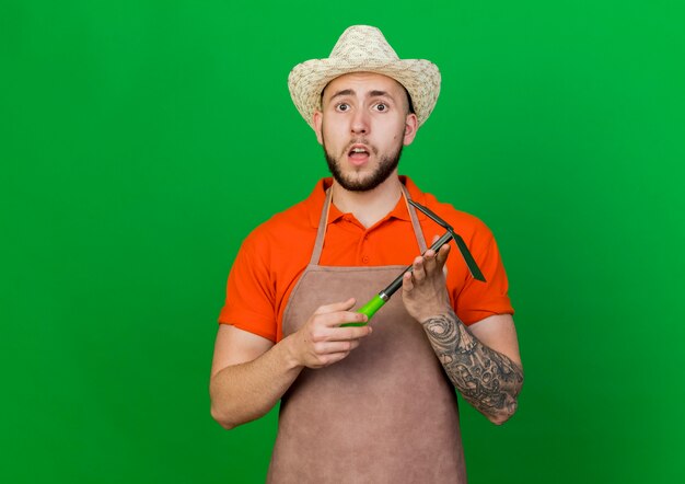 Surprised gardener man wearing gardening hat holds hoe rake