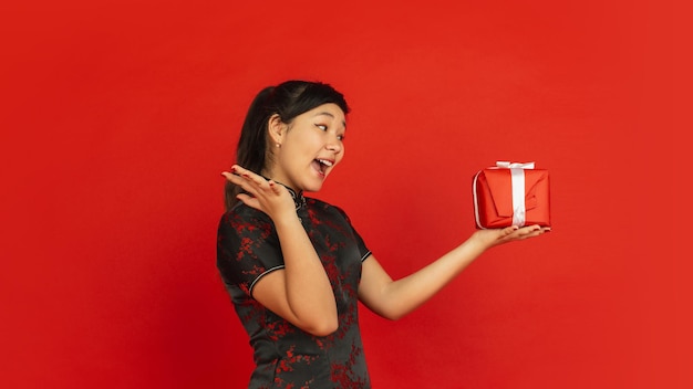 Surprised of gift. Happy Chinese New Year. Asian young girl's portrait isolated on red background. Female model in traditional clothes looks happy. Celebration, holiday, emotions. Copyspace.