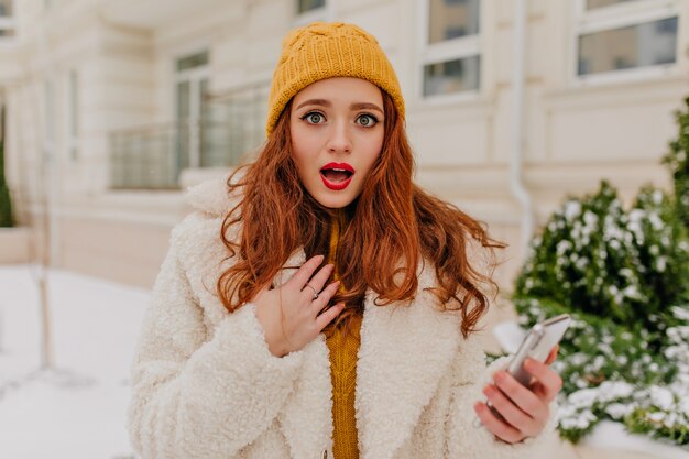 Удивленная рыжая девушка, стоящая на улице в холодный день. Великолепная молодая женщина с представлять телефона внешний.