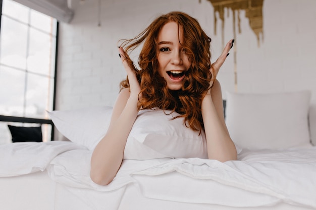 Ragazza sorpresa dello zenzero con la pelle pallida divertendosi la mattina presto. gioconda donna caucasica con capelli ricci scuri in posa in camera da letto.