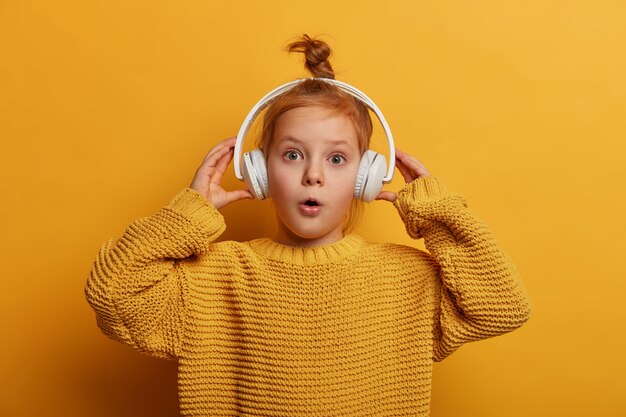 驚いた生姜の子供は、ヘッドフォンでオーディオトラックを聴き、大きな音に感銘を受け、驚異的に口を開き、黄色い壁に隔離された特大のニットセーターを着ています。子供と趣味の概念