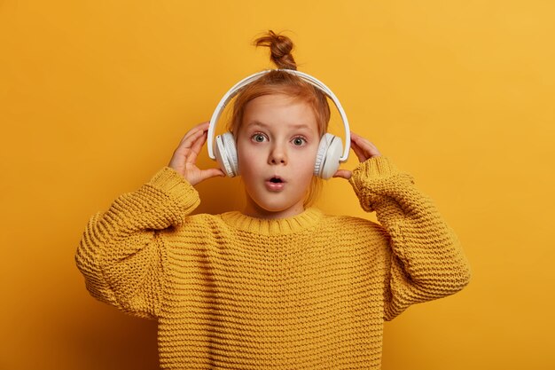 Удивленный рыжий ребенок слушает звуковую дорожку в наушниках, впечатлен громким звуком, удивленно открывает рот, носит большой вязаный свитер, изолированный на желтой стене. Дети и концепция хобби