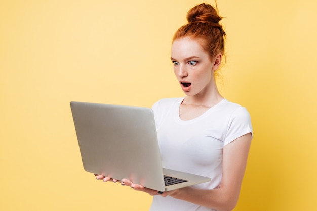 Удивленная рыжая женщина в футболке держит и смотрит на ноутбук