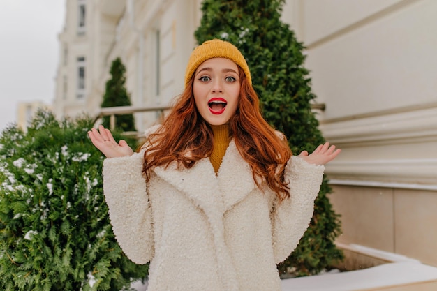 Surprised ginger woman in white coat waving hands on the street Outdoor photo of amazed girl in coat and hat