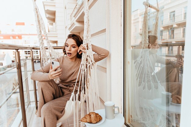 Удивленная девушка в коричневом свитере позирует с телефоном на балконе. Пораженная очаровательная дама обедает на террасе.