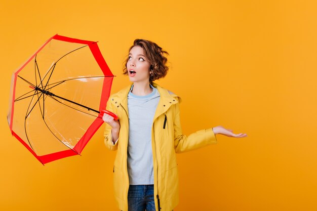 Surprised girl in coat looking up and holding umbrella.  shocked young lady with parasol isolated on bright orange wall.