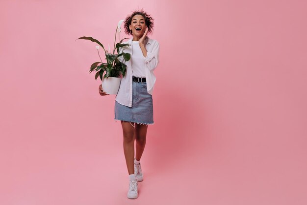 Surprised girl in denim skirt posing with pot on isolated background Portrait of woman in white shirt holding flower on pink backdrop