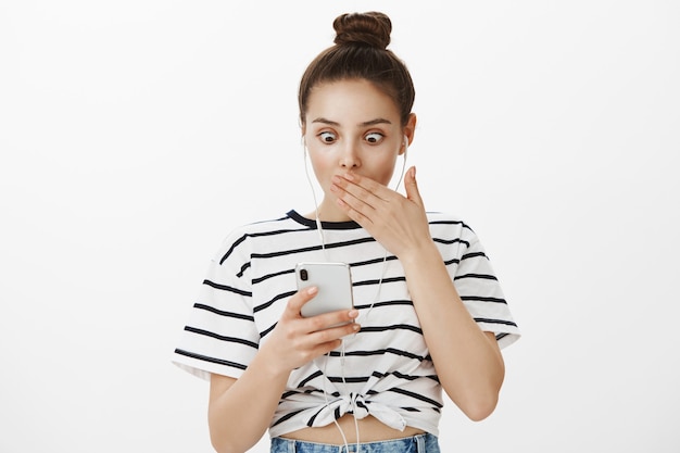 Surprised girl gasping, stare at smartphone with shocked face, wearing headphones