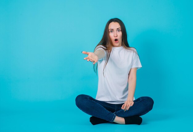 Surprised girl is extending hand to camera by sitting on floor on blue background