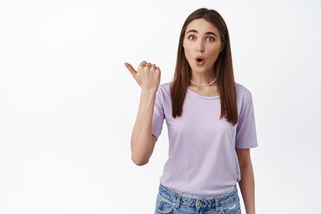 Surprised girl looks curious about advertisement beside her, pointing left and look amazed, asks question about event promo logo, showing banner, white background.