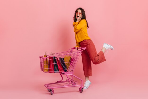 Surprised girl in pink culottes posing with trolley full of multi-colored packages with new clothes.