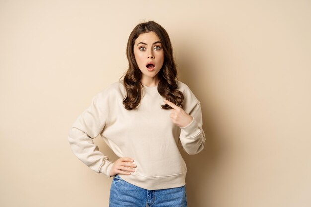 Surprised girl pointing at herself and look with disbelief standing over beige background