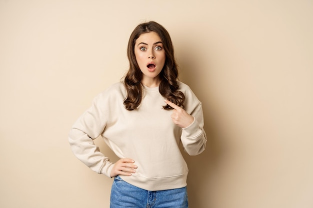 Surprised girl pointing at herself and look with disbelief standing over beige background