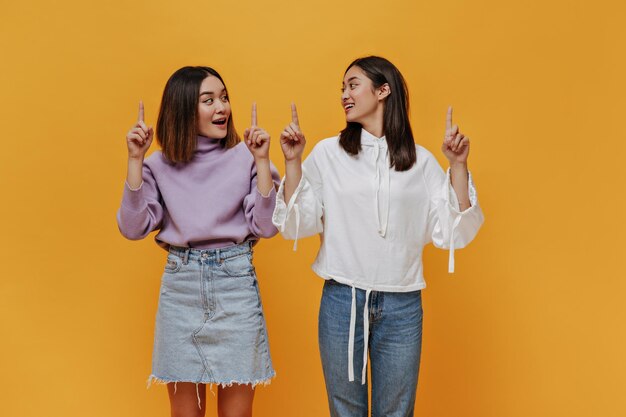 Surprised girl in purple sweater and denim skirt looks at friend Women in stylish outfits point up at place for text on isolated orange background