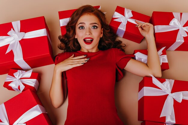 Surprised girl in red dress lying on the floor near presents. Pleased white female model having fun in birthday.