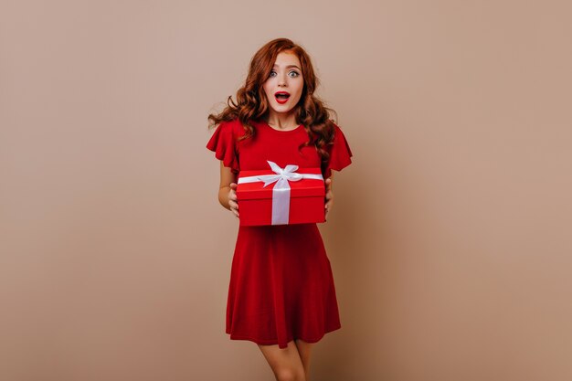 Surprised girl in short red dress holding gift. Adorable long-haired woman preparing new year presents.