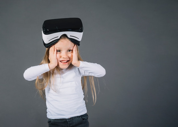 Surprised girl wearing virtual reality glasses on her head touching her cheeks against gray background