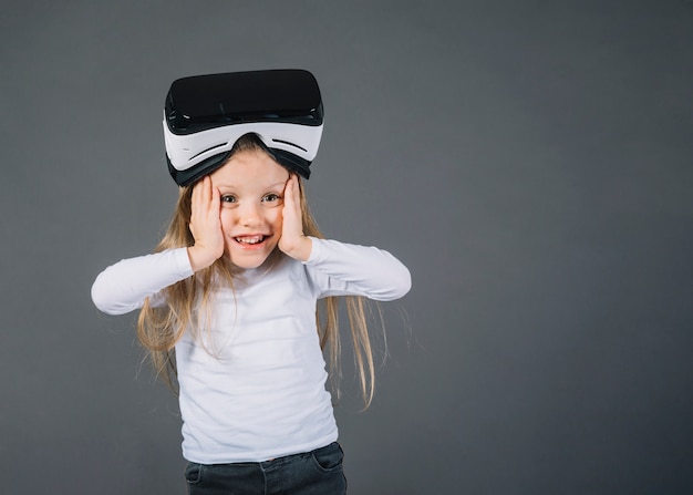 Бесплатное фото Удивленная девушка в очках виртуальной реальности на голове, касаясь щеками на сером фоне