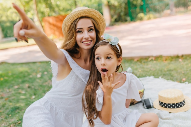 Surprised girl with big dark eyes looking where her mother pointing with finger. Charming young woman with long curly hair having fun with brunette cute daughter wears ribbon.