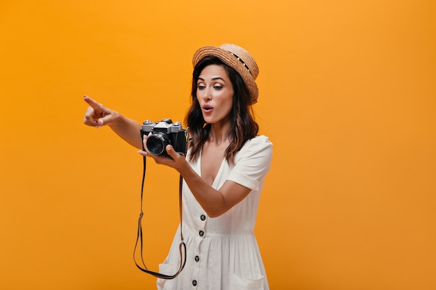 Surprised girl with retro camera shows finger into distance. Cute brunette with short hair in light stylish clothes posing.