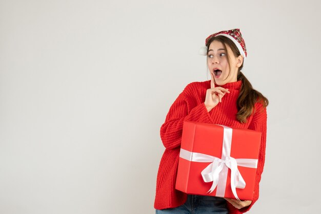 surprised girl with santa hat holding heavy gift standing on white