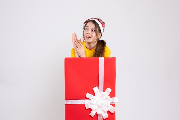 surprised girl with santa hat standing behind big xmas gift on white