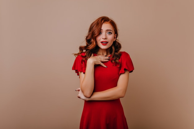 Surprised glamorous girl with curly hairstyle posing . Spectacular ginger lady in red outfit waiting for party.