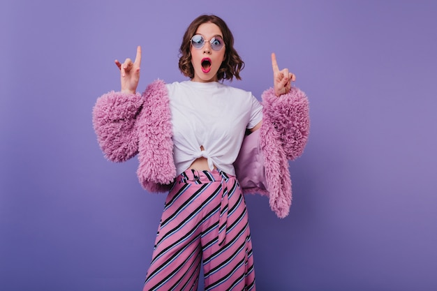 Surprised glamorous woman in striped pants posing on bright purple wall. Indoor portrait of emotional girl with wavy hair expressing amazement.
