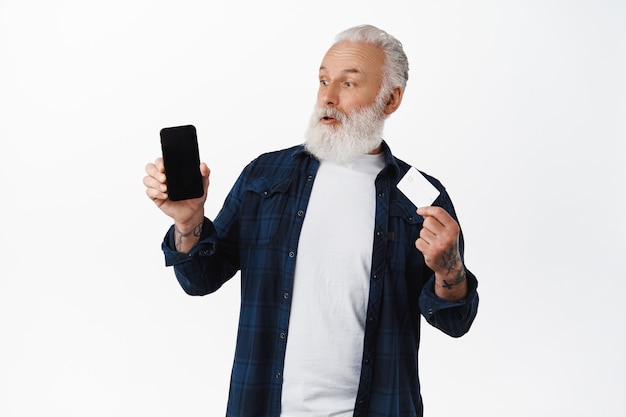 Удивленный дедушка смотрит на экран смартфона, показывая кредитную карту, изумленный покупками онлайн-приложения, стоящего у белой стены