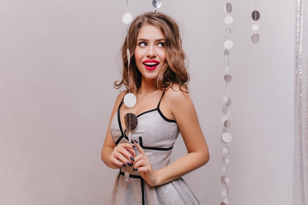 Surprised green-eyed girl mysteriously looks up and posing on gray background with sparkles. Portrait of woman with red lipstick in beautiful outfit