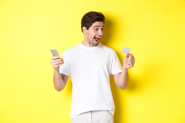 Surprised guy holding smartphone and credit card, online shopping on black friday, standing over yellow background.