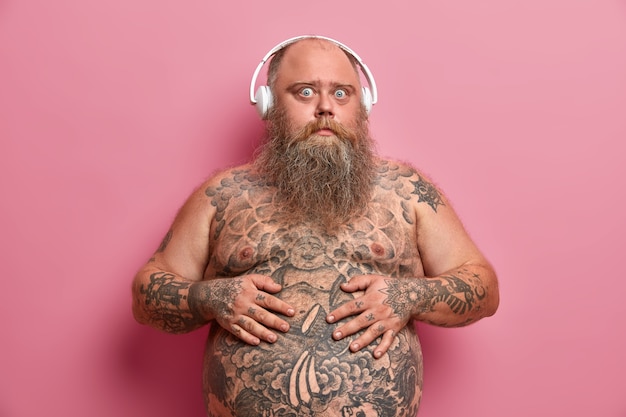 Ragazzo sorpreso tiene le mani sulla pancia, ha tatuato lo stomaco e le braccia, sta con il corpo nudo, essendo in sovrappeso, ascolta la traccia audio preferita in cuffia, isolato sul muro rosa.
