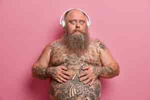 Free photo surprised guy keeps hands on tummy, has tattooed stomach and arms, stands with naked body, being overweight, listens favouite audio track in headphones, isolated on pink wall.