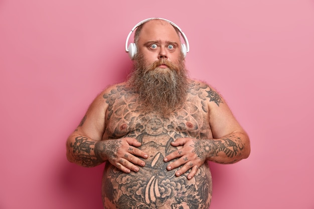  surprised guy keeps hands on tummy, has tattooed stomach and arms, stands with naked body, being overweight, listens favouite audio track in headphones, isolated on pink wall.