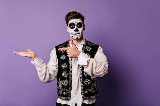 Surprised guy in national Mexican vest pointing finger to left. Portrait of man with painted face with place for tex on lilac wall.