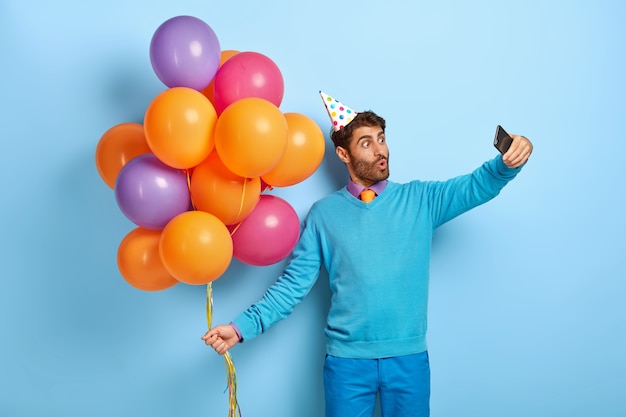 Surprised guy with birthday hat and balloons posing in blue sweater