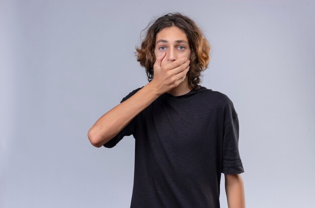 Surprised guy with long hair in black t-shirt covered his mouth with hand on white background