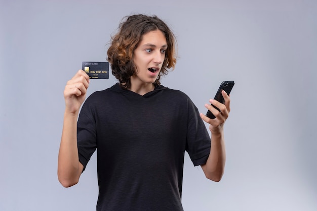 白い背景の上の電話と銀行カードを保持している黒のtシャツに長い髪の驚いた男