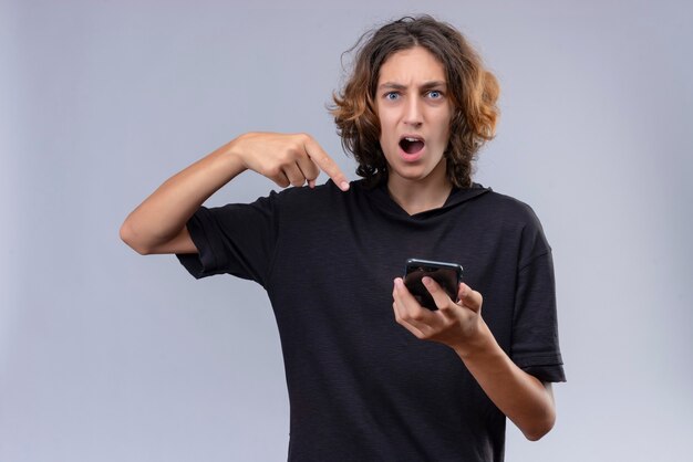 Surprised guy with long hair in black t-shirt holding a phone and points to phone on white background