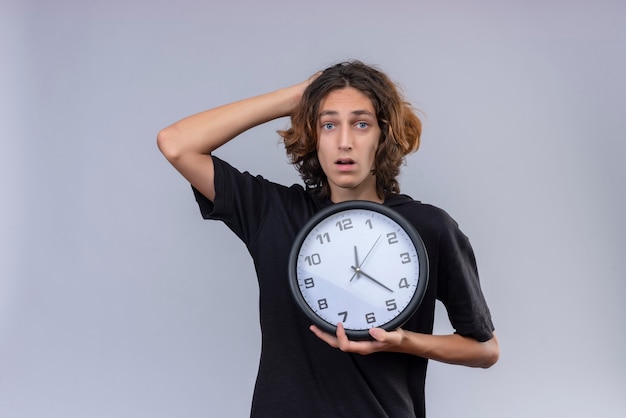 Удивленный парень с длинными волосами в черной футболке держит настенные часы и схватился за голову на белом фоне