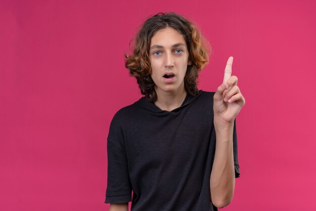 Surprised guy with long hair in black t-shirt points to up on pink background