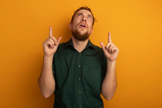 Surprised handsome blonde man looks and points up with two hands isolated on orange wall