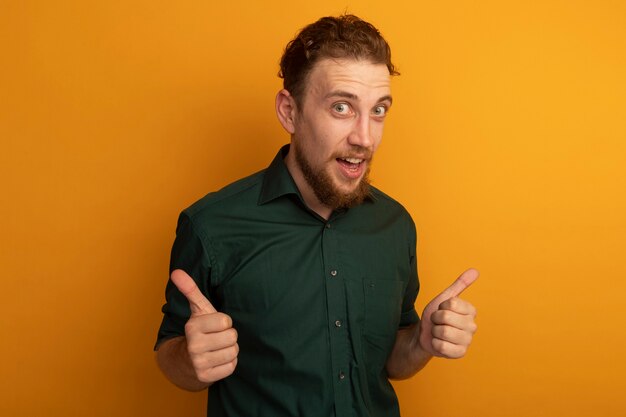 Удивленный красивый белокурый мужчина показывает палец вверх двух рук, изолированных на оранжевой стене