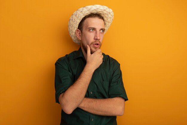 ビーチ帽子をかぶった驚いたハンサムなブロンドの男は、あごに手を置き、オレンジ色の壁に隔離された側を見て