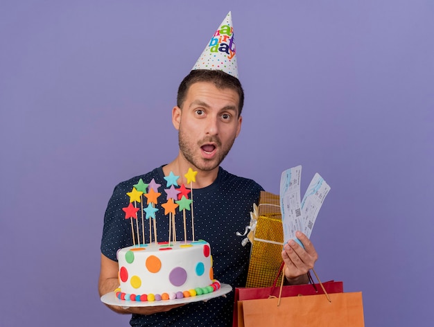 Berretto da portare di compleanno dell'uomo caucasico bello sorpreso tiene il contenitore di regalo del sacchetto della spesa di carta della torta di compleanno e biglietti aerei isolati su fondo viola con lo spazio della copia