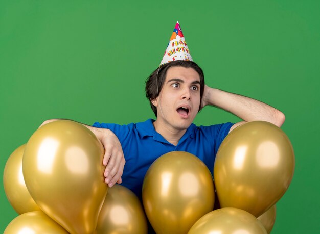 Surprised handsome caucasian man wearing birthday cap puts hand on head behind standing with helium balloons 