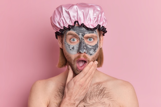 surprised handsome guy applies beauty face mask keeps mouth opened wears bath hat has naked body.