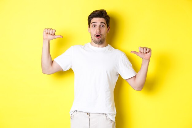 Surprised handsome guy pointing at himself, looking amazed, standing over yellow background