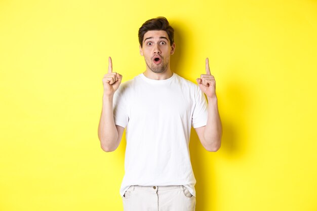 Surprised handsome guy in white t-shirt, pointing fingers up, interested about advertisement, standing against yellow background.